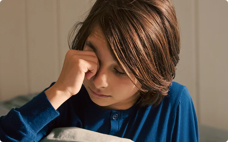En gutt sitter i sengen sin og klør seg i øyet. Husstøvmidd trives i og rundt sengen og kan særlig forårsake symptomer om natten og morgenen.